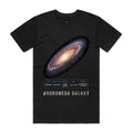 Front of Voltare Andromeda Galaxy black tee with a glowing cream-gold core, blue-violet spiral arms, galaxy-facts panel and silver-grey lettering