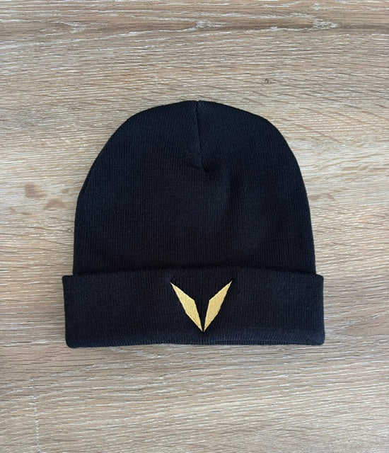 Actual black Voltare knit beanie sample on a wooden table, showing the folded cuff with solid gold V embroidery.