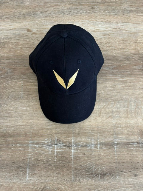 Actual black Voltare cap sample on a wooden table, showing the solid gold V embroidery on the front panel.