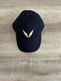 Actual black Voltare cap sample on a wooden table, showing the solid gold V embroidery on the front panel.